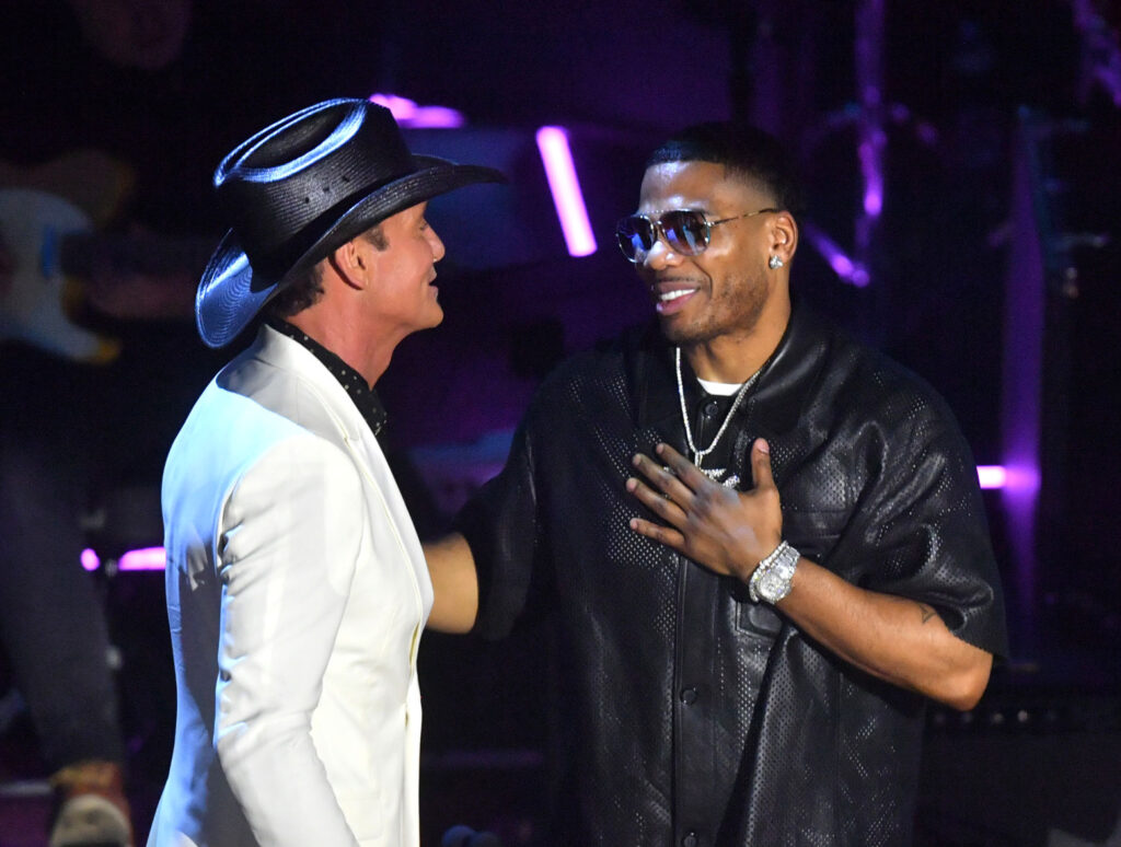 Tim McGraw is on stage in a white suit and cowboy hat, with Nelly dressed in black and sunglasses.